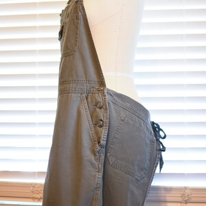 May include: A pair of olive green denim overalls with a single front pocket and a button closure. The overalls have adjustable straps and a tie at the back.