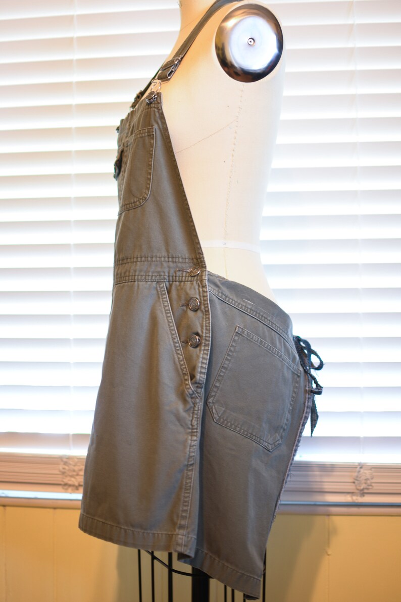 May include: A pair of olive green denim overalls with a single front pocket and a button closure. The overalls have adjustable straps and a tie at the back.