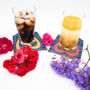 May include: Two glasses of beverages, one dark and one light, sit on denim coasters with colorful designs. Red and pink roses and purple flowers are arranged around the glasses, creating a vibrant and inviting scene.