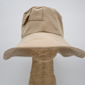 Puede incluir: Un sombrero de pescador beige con ala ancha, exhibido en una forma de cabeza tejida. El sombrero tiene una corona estructurada con un pequeño detalle de bolsillo y un ala suavemente curvada, que ofrece protección solar. Su color neutro es versátil.