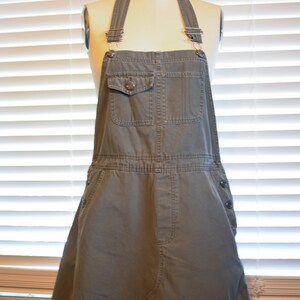 May include: A gray denim overall dress with a single button on the front pocket. The dress has adjustable straps and a skirt-like bottom.