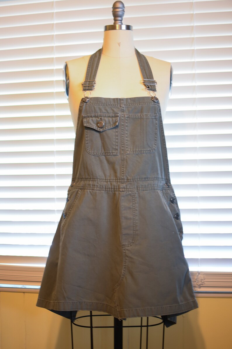 May include: A gray denim overall dress with a single button on the front pocket. The dress has adjustable straps and a skirt-like bottom.