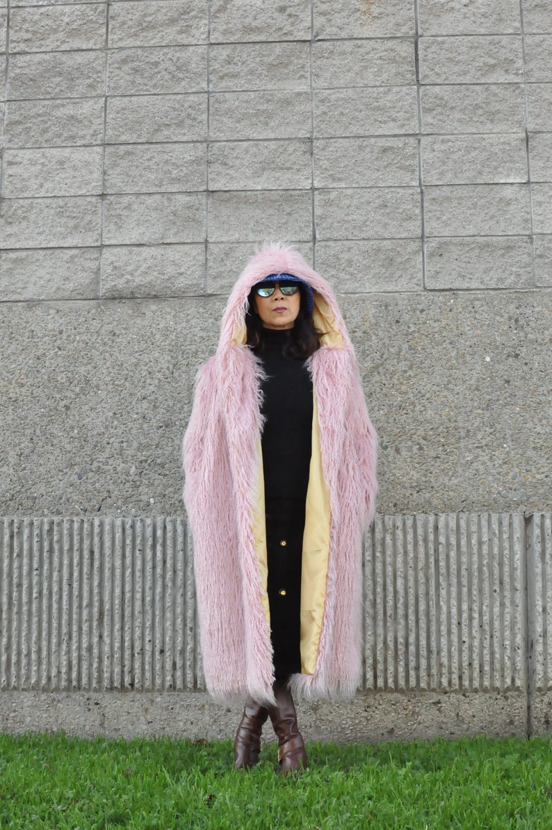 Puede incluir: Una chaqueta de piel sint&eacute;tica rosa con capucha y forro amarillo. La chaqueta la lleva puesta una persona con gafas de sol y botas marrones.