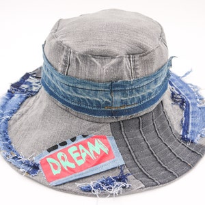 May include: A gray denim bucket hat with a blue denim band and frayed edges. The hat has a patch with the word "DREAM" in green letters on a red background.