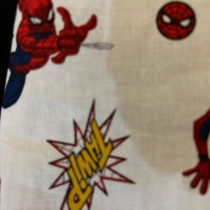 May include: White fabric with a red and blue Spiderman design. The fabric has a repeated pattern of Spiderman in different poses, including a Spiderman head and a speech bubble that says "THWIP".