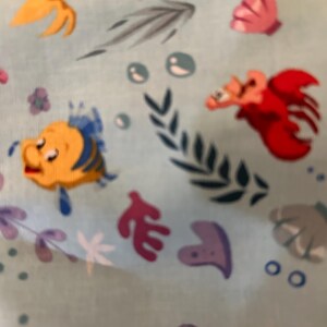 K&ouml;nnte beinhalten: Ein blauer Stoff mit einem sich wiederholenden Muster aus Illustrationen zum Thema Meer. Das Muster enth&auml;lt einen gelben Fisch mit einer blauen Flosse, einen roten Krebs, eine violette Muschel, eine rosa Muschel, einen gelben Seestern, eine violette Musiknote und gr&uuml;ne Algen.