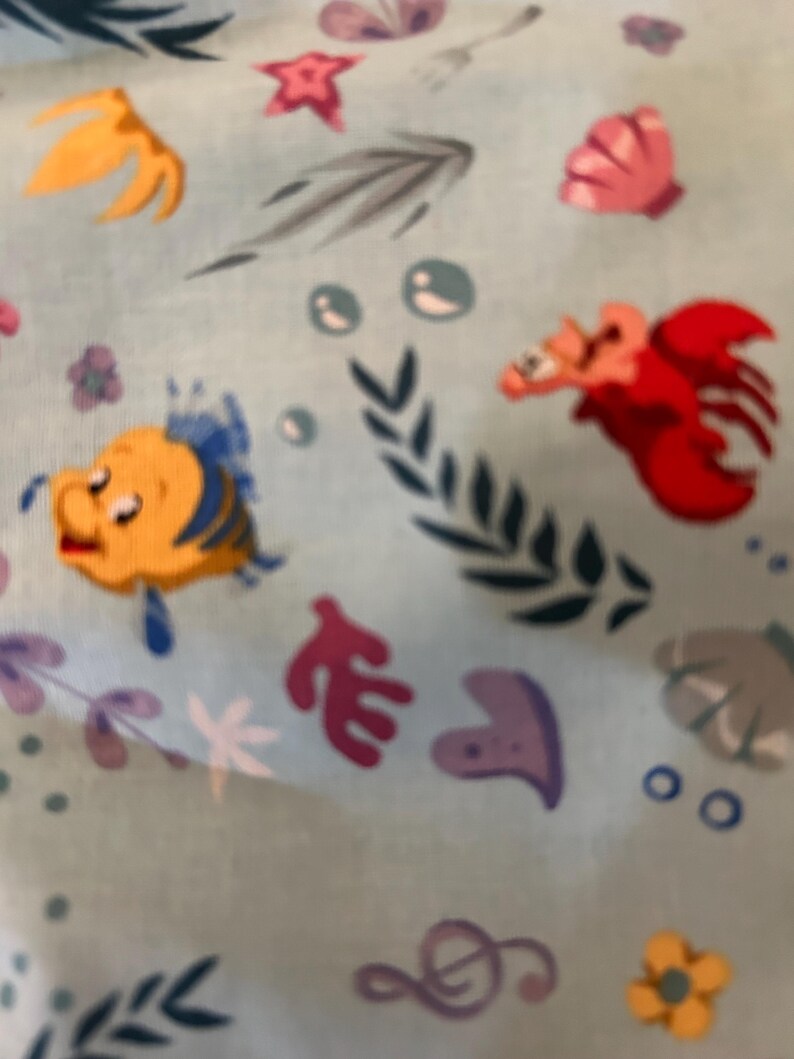 K&ouml;nnte beinhalten: Ein blauer Stoff mit einem sich wiederholenden Muster aus Illustrationen zum Thema Meer. Das Muster enth&auml;lt einen gelben Fisch mit einer blauen Flosse, einen roten Krebs, eine violette Muschel, eine rosa Muschel, einen gelben Seestern, eine violette Musiknote und gr&uuml;ne Algen.