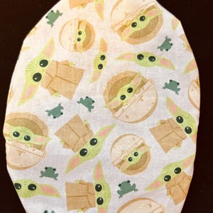 May include: White fabric item with a repeating pattern of a green alien character with large eyes, and small green frog-like creatures. The alien is depicted in various poses, some in a pod-like structure. The item has a rounded shape.