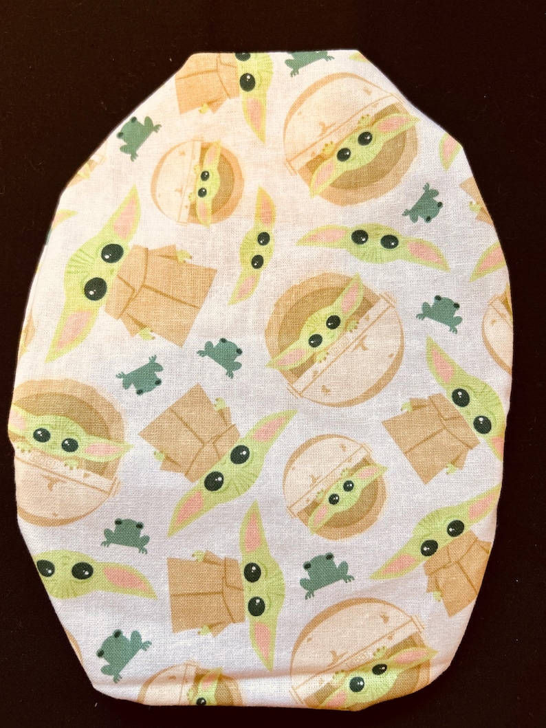 May include: White fabric item with a repeating pattern of a green alien character with large eyes, and small green frog-like creatures. The alien is depicted in various poses, some in a pod-like structure. The item has a rounded shape.