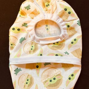 May include: A pet accessory featuring a repeating pattern of a green alien character and small green frogs on a white background. The accessory has a circular opening and a white elastic band.