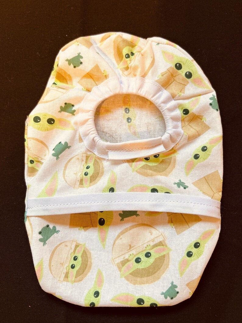 May include: A pet accessory featuring a repeating pattern of a green alien character and small green frogs on a white background. The accessory has a circular opening and a white elastic band.