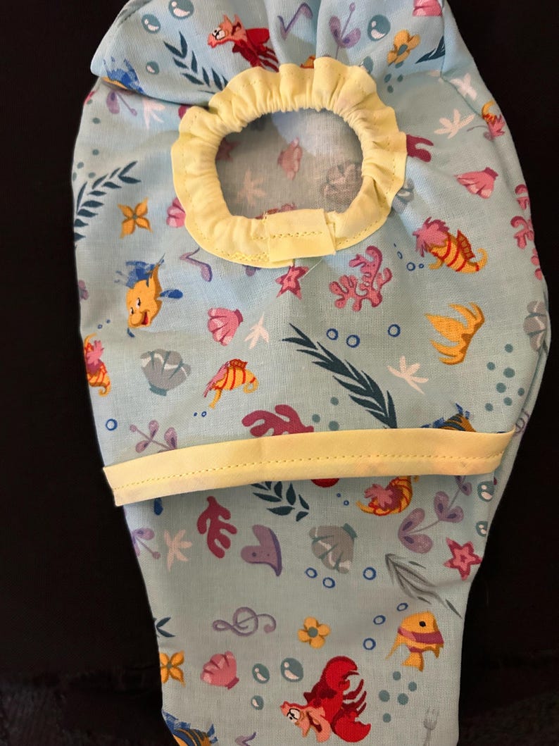 K&ouml;nnte beinhalten: Eine hellblaue Stofftasche mit gelbem Besatz und einem Kordelzug. Die Tasche hat ein sich wiederholendes Muster aus Meeresbewohnern, darunter Fische, Muscheln und Algen.