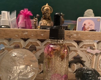 May include: A clear glass dropper bottle filled with a liquid containing silver glitter and dried pink flower petals. The bottle features a black dropper cap and is positioned between a clear spherical crystal and a dark, textured stone.