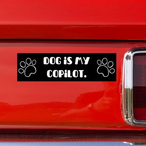 May include: Black rectangular bumper sticker with white text and paw prints. The text reads "DOG IS MY COPILOT." The sticker is attached to a red vehicle.