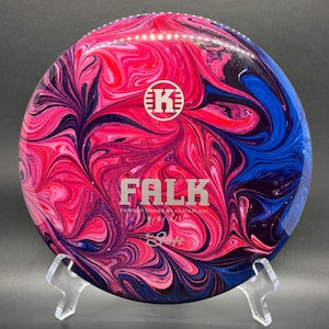 May include: A disc golf disc with a vibrant, swirled pattern in shades of pink, red, blue, and purple. The disc features the word "FALK" in silver lettering, along with the Kastaplast logo and other text. The disc is displayed on a clear stand.