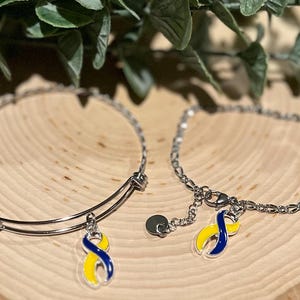 May include: Two silver bracelets with charms. One is a bangle style, the other a chain. Each charm is a blue and yellow ribbon, a symbol of Down syndrome awareness. The bracelets are displayed on a wooden surface.