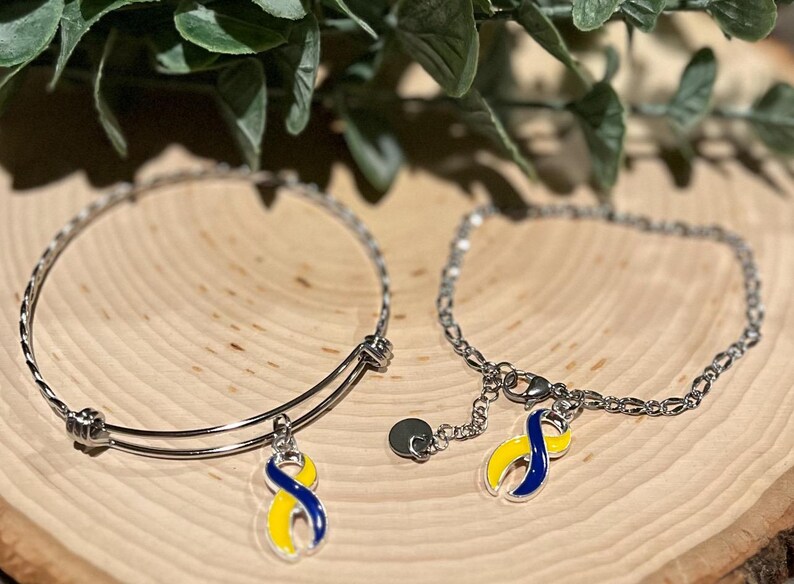 May include: Two silver bracelets with charms. One is a bangle style, the other a chain. Each charm is a blue and yellow ribbon, a symbol of Down syndrome awareness. The bracelets are displayed on a wooden surface.