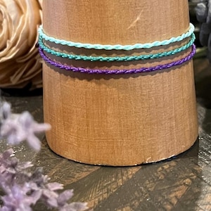 Pode incluir: Três pulseiras de amizade trançadas em tons de roxo, azul-petróleo e azul claro, exibidas em uma superfície de madeira.
