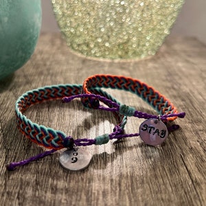 May include: Two braided friendship bracelets with teal, orange, and purple threads. Each bracelet has a small, round metal charm. One charm has the number "9" and the other says "STAY".