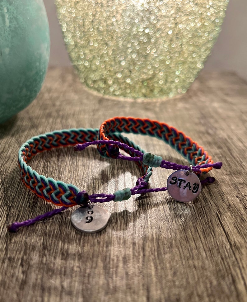 May include: Two braided friendship bracelets with teal, orange, and purple threads. Each bracelet has a small, round metal charm. One charm has the number "9" and the other says "STAY".