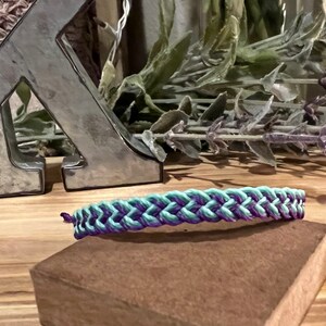 May include: A braided bracelet in shades of teal and purple rests on a small, brown block. The bracelet is the focal point, with a woven design. The background includes a silver letter and dried lavender.