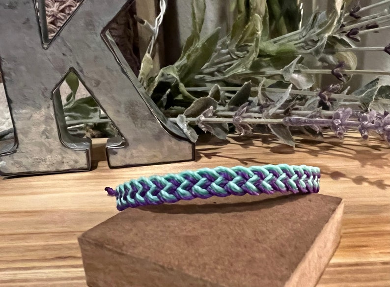 May include: A braided bracelet in shades of teal and purple rests on a small, brown block. The bracelet is the focal point, with a woven design. The background includes a silver letter and dried lavender.
