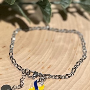 May include: Silver-toned chain bracelet with a charm featuring a yellow and blue ribbon design. The bracelet has a lobster clasp closure and a small, round charm. The ribbon charm is a symbol of awareness.