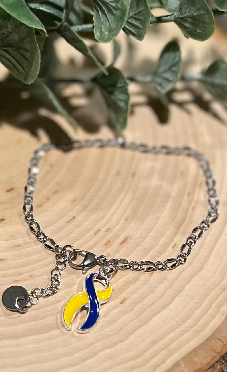 May include: Silver-toned chain bracelet with a charm featuring a yellow and blue ribbon design. The bracelet has a lobster clasp closure and a small, round charm. The ribbon charm is a symbol of awareness.