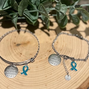 Puede incluir: Dos pulseras plateadas con dijes. Una es un brazalete, la otra una cadena. Cada una tiene un dije redondo con el texto "She believed she could so she did" y un dije de cinta turquesa. Las pulseras se exhiben sobre una superficie de madera.