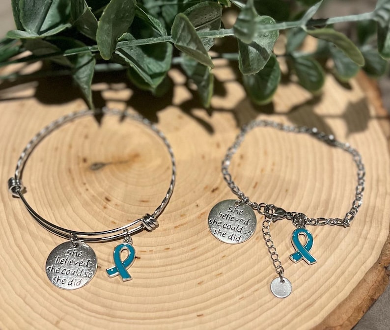 Puede incluir: Dos pulseras plateadas con dijes. Una es un brazalete, la otra una cadena. Cada una tiene un dije redondo con el texto "She believed she could so she did" y un dije de cinta turquesa. Las pulseras se exhiben sobre una superficie de madera.