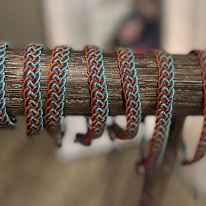 May include: Braided bracelets in teal, orange, and navy blue are displayed on a wooden dowel. The bracelets have a woven design, with the colors alternating in a repeating pattern. The bracelets are a handmade accessory.