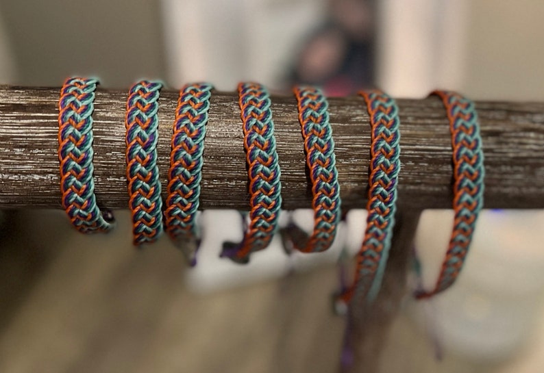 May include: Braided bracelets in teal, orange, and navy blue are displayed on a wooden dowel. The bracelets have a woven design, with the colors alternating in a repeating pattern. The bracelets are a handmade accessory.