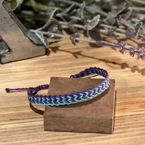 Pode incluir: Pulseira trançada em tons de roxo e verde-azulado. A pulseira é feita de fios tecidos, com fecho de nó deslizante. É exibida em um pequeno bloco retangular marrom, com fundo de madeira e raminhos de lavanda.
