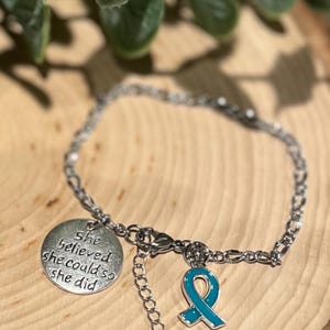Puede incluir: Pulsera plateada con un dije redondo con la inscripci&oacute;n "She believed she could so she did". Un dije de lazo turquesa y un peque&ntilde;o dije redondo cuelgan de la pulsera. La pulsera est&aacute; sobre una superficie de madera.