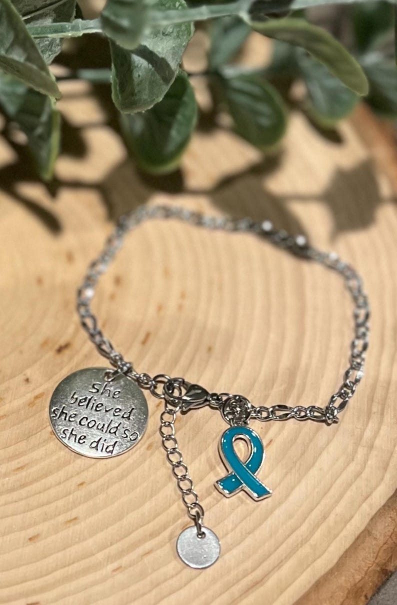 Puede incluir: Pulsera plateada con un dije redondo con la inscripci&oacute;n "She believed she could so she did". Un dije de lazo turquesa y un peque&ntilde;o dije redondo cuelgan de la pulsera. La pulsera est&aacute; sobre una superficie de madera.