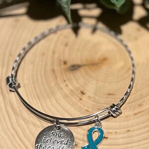 Puede incluir: Pulsera de brazalete plateada con dos colgantes. Un colgante es un disco redondo con la inscripci&oacute;n "She believed she could so she did". El otro es un colgante de cinta turquesa. La pulsera se exhibe sobre una superficie de madera.