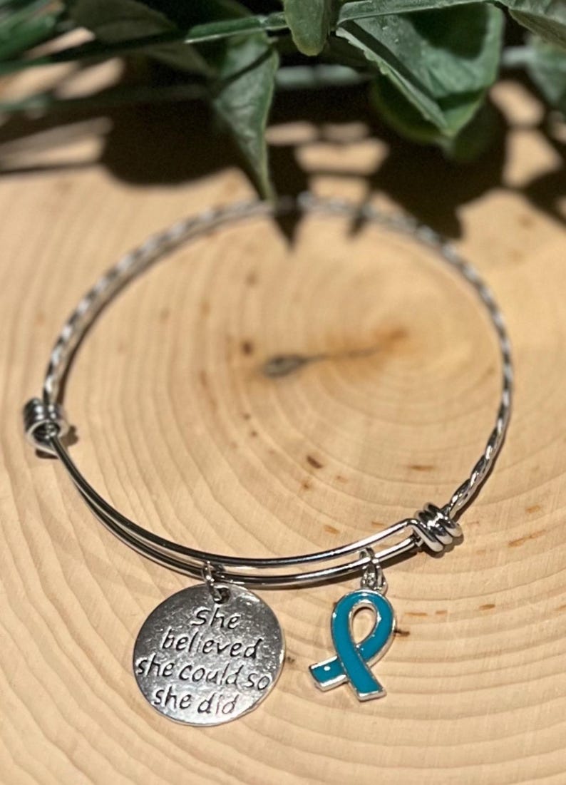 Puede incluir: Pulsera de brazalete plateada con dos colgantes. Un colgante es un disco redondo con la inscripci&oacute;n "She believed she could so she did". El otro es un colgante de cinta turquesa. La pulsera se exhibe sobre una superficie de madera.