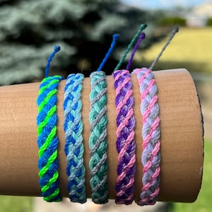 May include: Six colorful braided friendship bracelets in shades of blue, green, purple, and pink. Each bracelet is approximately 0.5 inches wide. The bracelets are displayed on a brown cylindrical object against a blurred green background.