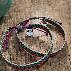 May include: Two braided bracelets with a mix of teal, orange, and purple threads. Each bracelet has a small silver charm with the word "STAY" stamped on it. The bracelets are on a wooden surface.