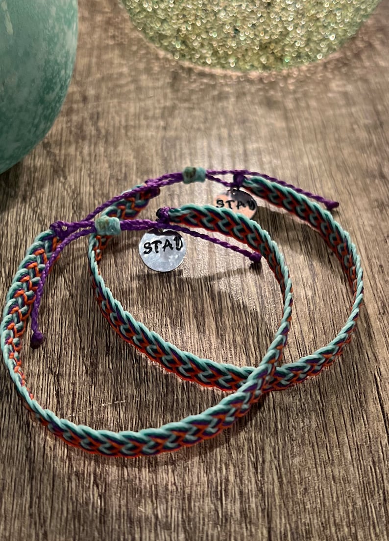 May include: Two braided bracelets with a mix of teal, orange, and purple threads. Each bracelet has a small silver charm with the word "STAY" stamped on it. The bracelets are on a wooden surface.