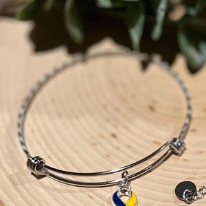 May include: A silver-colored bangle bracelet with a charm featuring a blue and yellow ribbon design. The charm is a symbol of awareness. The bracelet is displayed on a wooden surface.