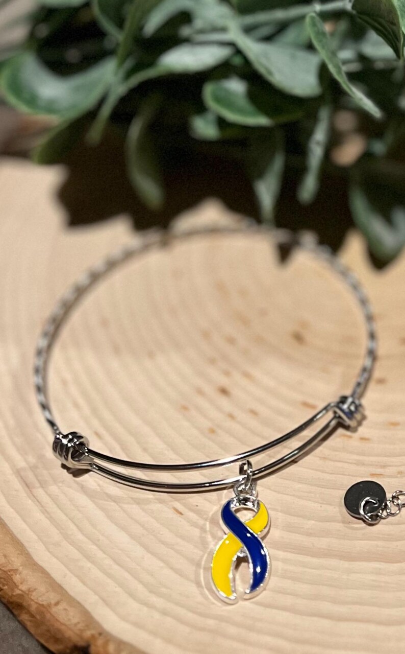 May include: A silver-colored bangle bracelet with a charm featuring a blue and yellow ribbon design. The charm is a symbol of awareness. The bracelet is displayed on a wooden surface.