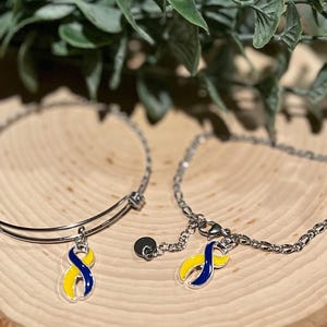 May include: Two silver bracelets with charms. One is a bangle style, the other a chain. Each charm is a yellow and blue ribbon, a symbol of Down syndrome awareness. The bracelets rest on a wooden surface with green foliage in the background.