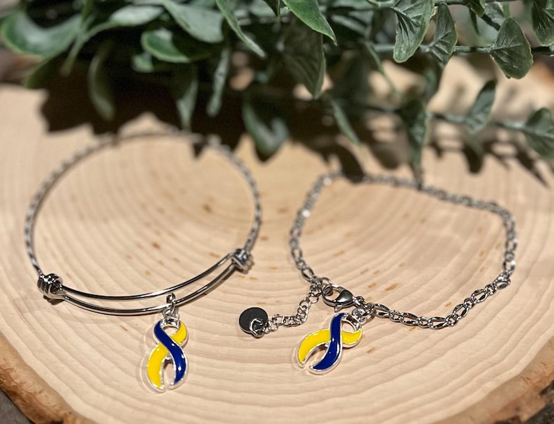 May include: Two silver bracelets with charms. One is a bangle style, the other a chain. Each charm is a yellow and blue ribbon, a symbol of Down syndrome awareness. The bracelets rest on a wooden surface with green foliage in the background.