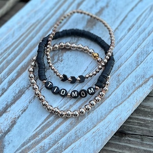 K&ouml;nnte beinhalten: Drei Armb&auml;nder, die auf einer Holzoberfl&auml;che gestapelt sind. Das untere Armband besteht aus goldenen Perlen und buchstabiert "TID MOM". Das mittlere Armband besteht aus schwarzen Perlen mit Pfeil-Anh&auml;ngern. Das obere Armband besteht aus schwarzen Perlen.