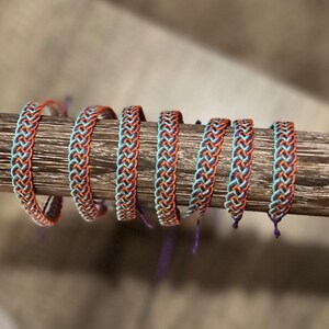 May include: Braided bracelets in a row, featuring a repeating pattern of orange, teal, and navy blue threads. The bracelets are displayed on a wooden dowel, showcasing their woven texture and color combinations. The bracelets are handmade.