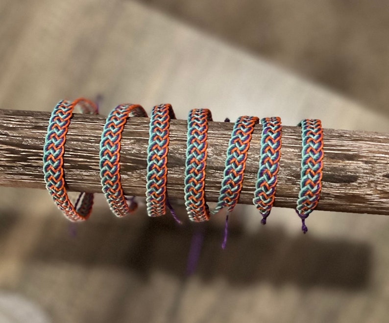 May include: Braided bracelets in a row, featuring a repeating pattern of orange, teal, and navy blue threads. The bracelets are displayed on a wooden dowel, showcasing their woven texture and color combinations. The bracelets are handmade.