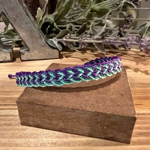 May include: Braided bracelet in shades of purple and mint green. The bracelet is displayed on a small, brown, rectangular block. The background includes decorative elements and a wooden surface.