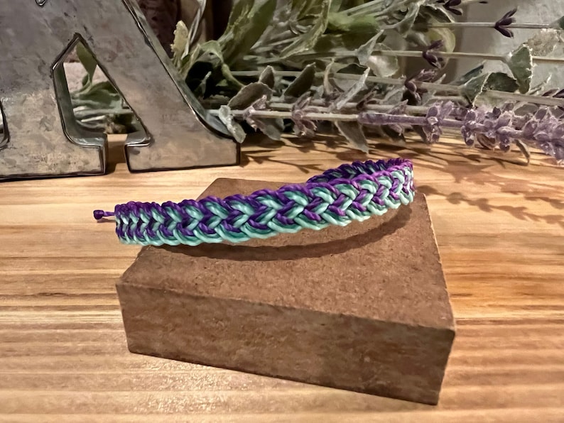 May include: Braided bracelet in shades of purple and mint green. The bracelet is displayed on a small, brown, rectangular block. The background includes decorative elements and a wooden surface.
