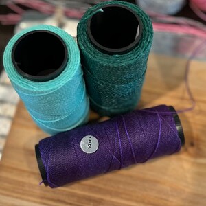 May include: Three spools of thread in shades of teal, green, and purple. The purple spool has a small, round tag with the number 9 on it. The spools are arranged on a wooden surface, with a blurred background.